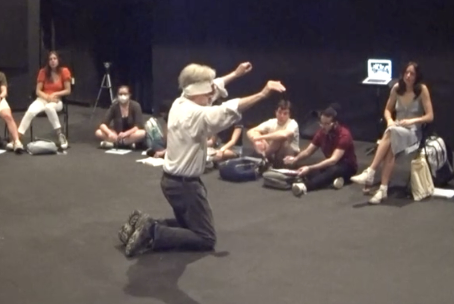 Dan Lloyd on his knees on a black stage, arms raised, surrounded by seated students